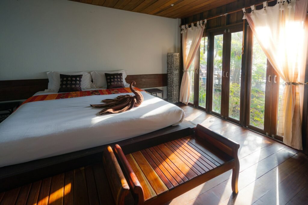Sunlit hotel room with a low white bed, wooden bench, and tall windows opening onto greenery